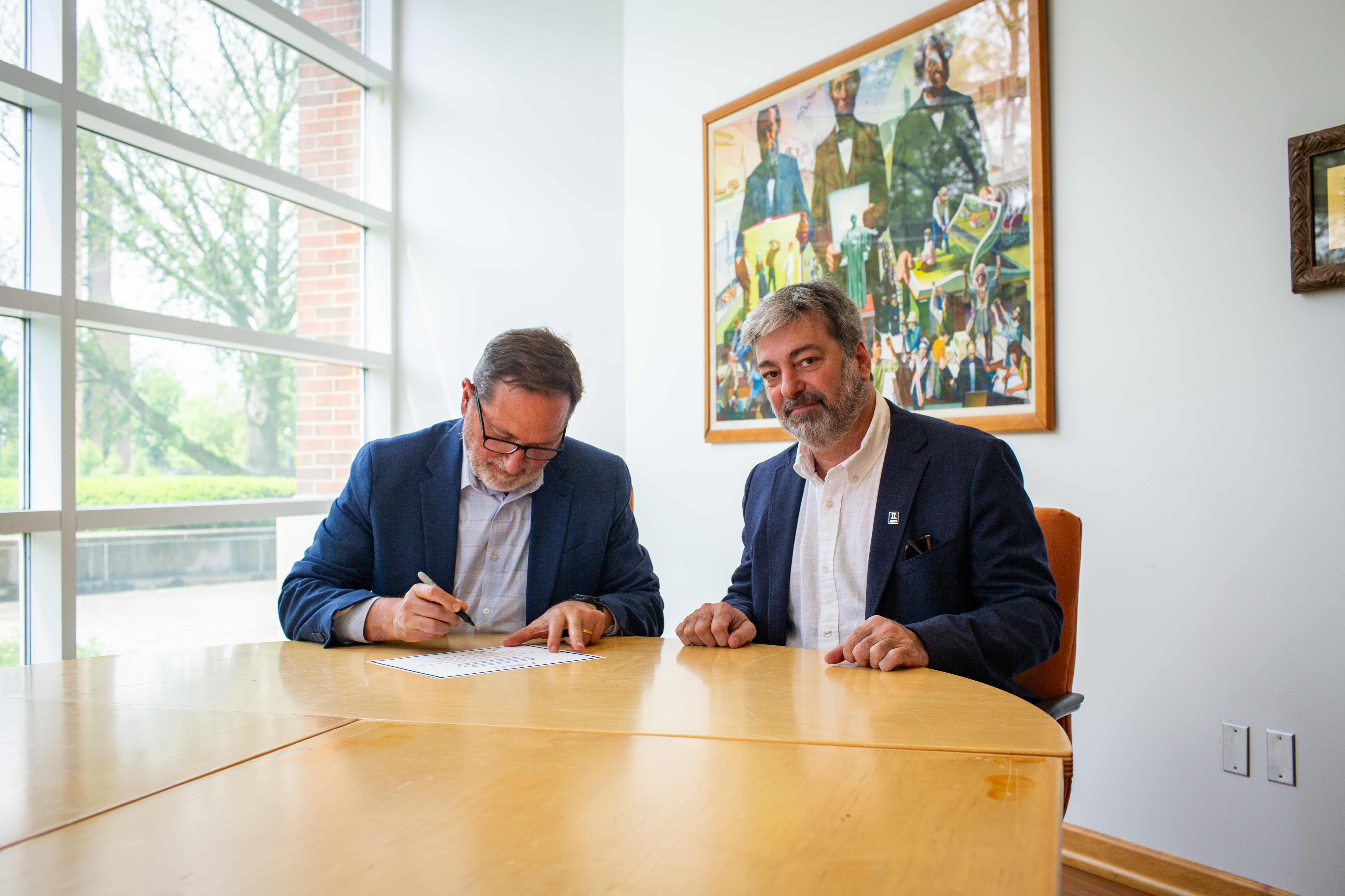 Two men sit at a table signing a document. 
